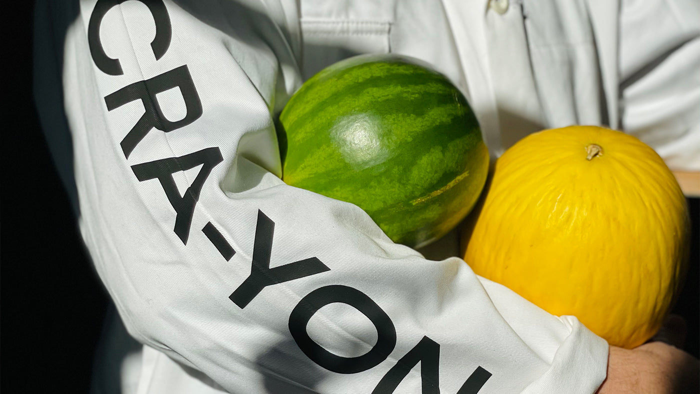 Person in CRA-YON jacket holding a watermelon and a yellow melon in bright natural light.
