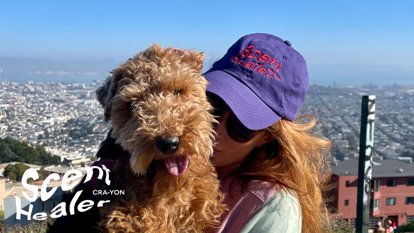 Person in purple cap cuddling fluffy dog with city view in background and 'Scent Healer' CRA-YON logo overlay