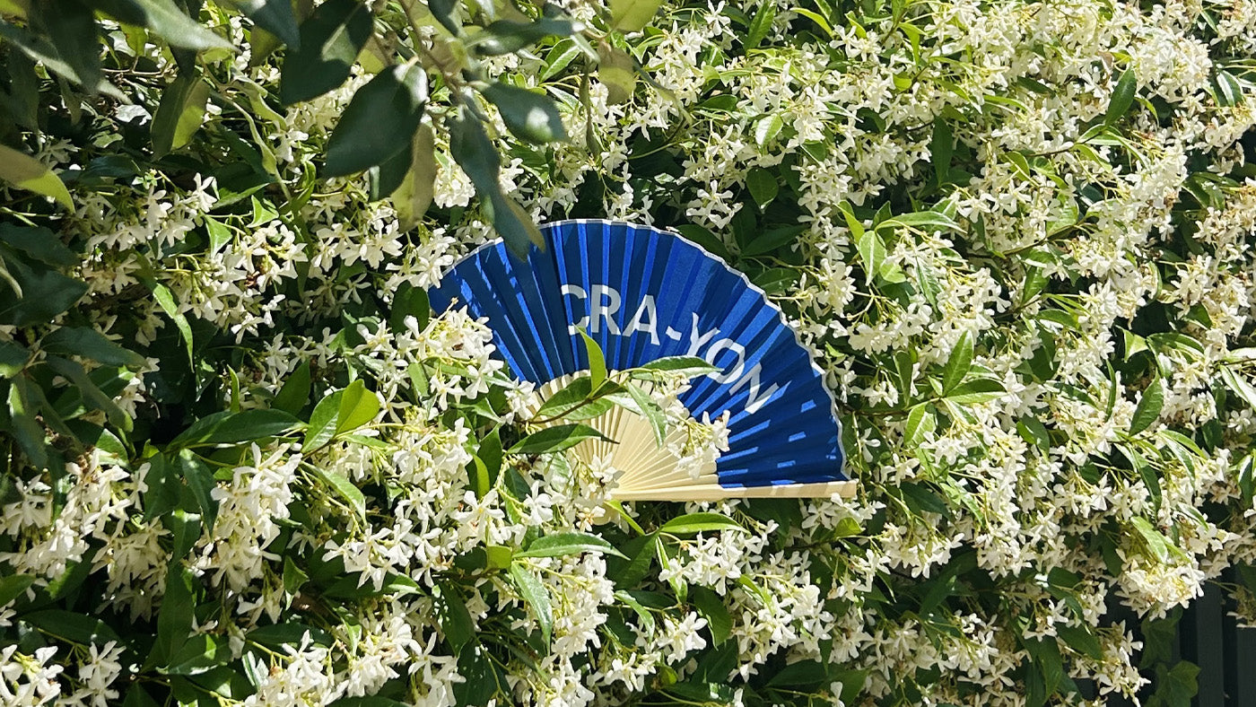 CRA-YON blue fan placed among blooming white jasmine flowers in lush greenery