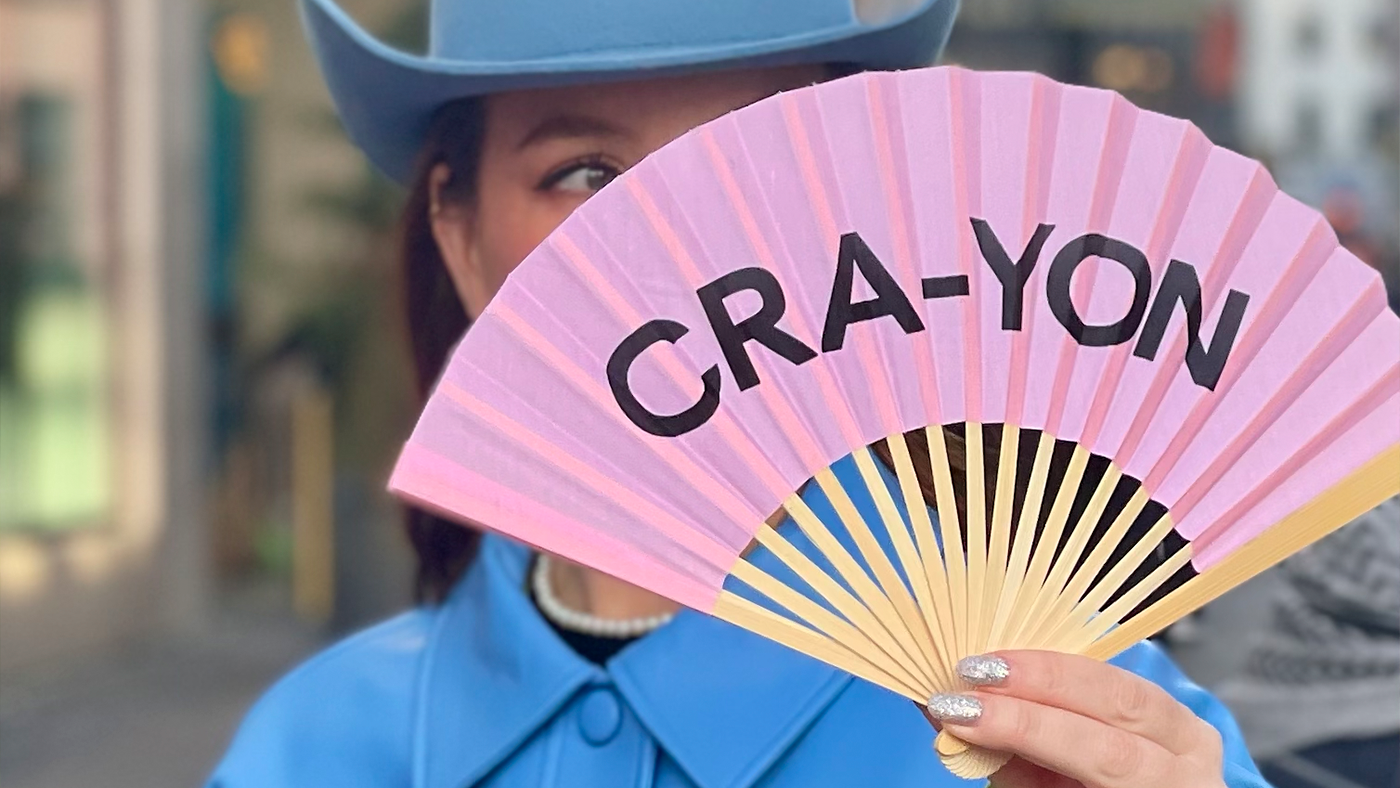 Person in blue coat and hat holding pink CRA-YON branded fan in front of face on city street