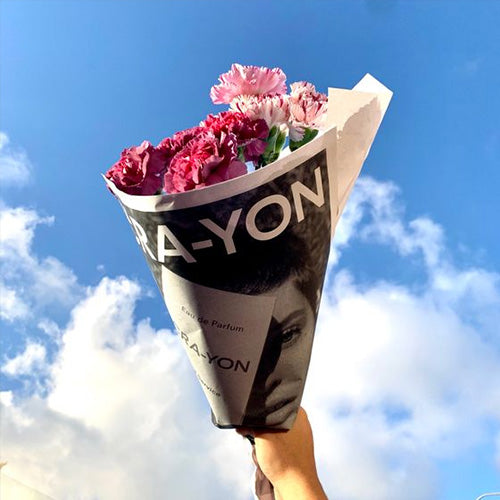 Hand holding a flower bouquet wrapped in CRA-YON branded paper against a bright blue sky with clouds