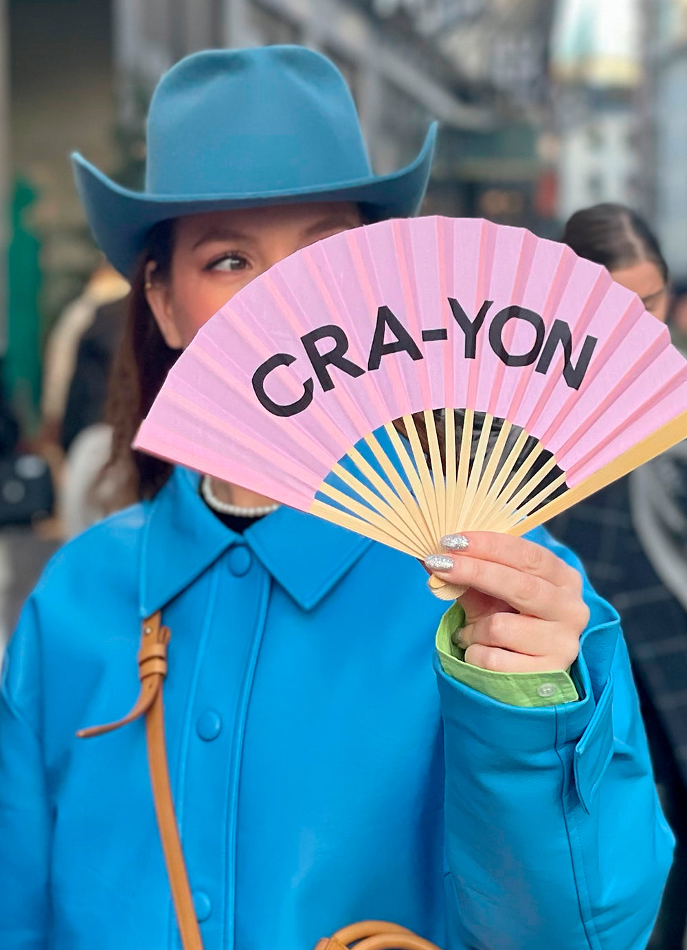 Fashion-forward woman in blue coat and cowboy hat holding pink CRA-YON fan in vibrant city street scene