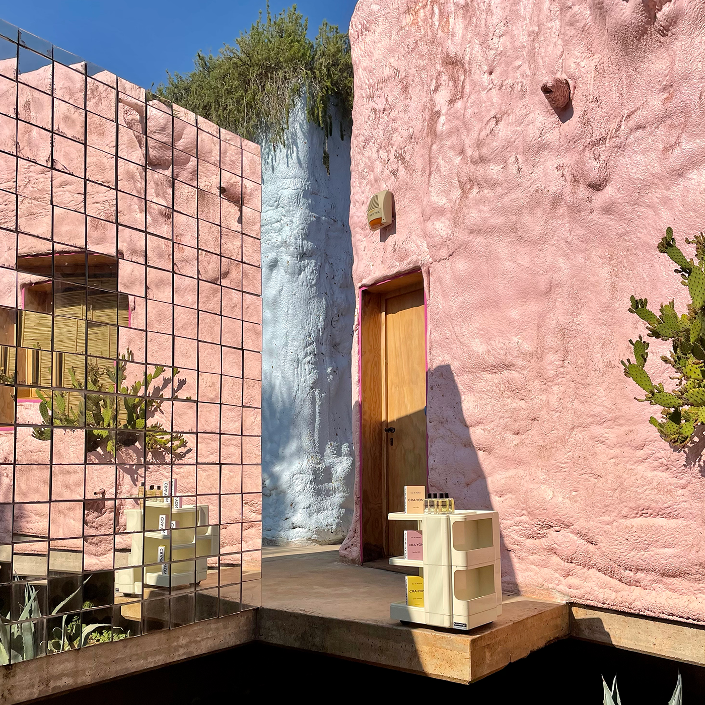 CRA-YON perfumes in a pink desert-style setup with cactus, mirror wall, and natural sunlight