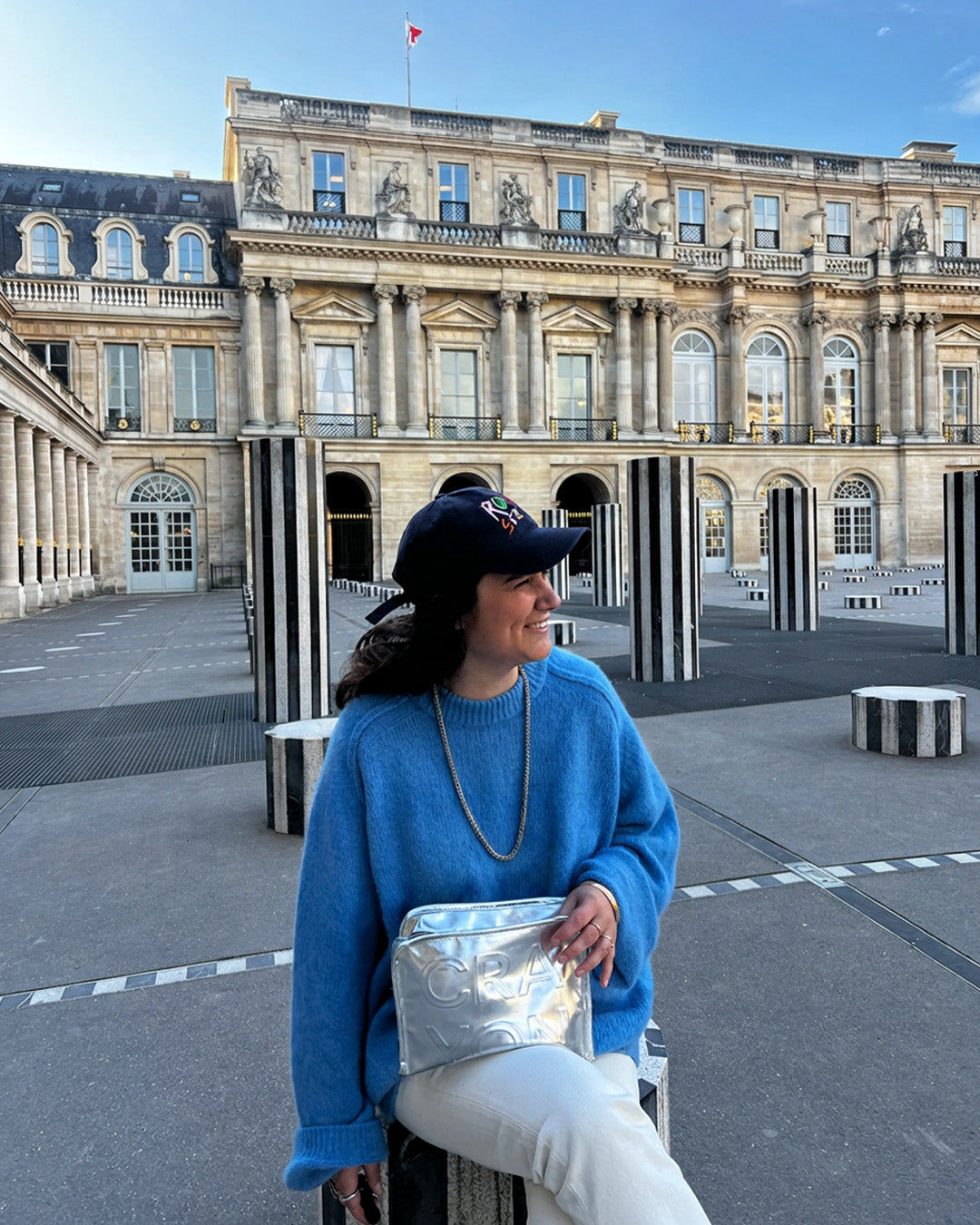 Christine, co-founder of CRA-YON, at Palais Royal in Paris wearing a blue sweater and CRA-YON cap, holding a silver CRA-YON bag.