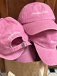 Pink embroidered caps reading 'CRA-YON' and 'Vanilla CEO' stacked in a brown paper bag against wooden background