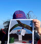 Person with a purple CRA-YON cap holding a poster with a desert scene and product display against a blue sky.
