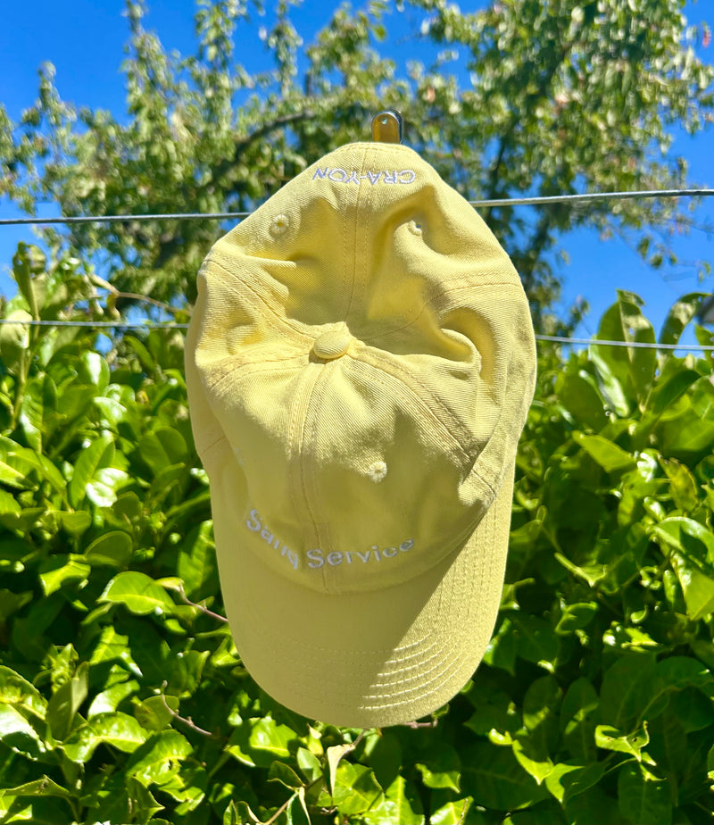 Light yellow cap with embroidered text ‘Sand Service’, hanging on a line against a bright green hedge and clear blue sky
