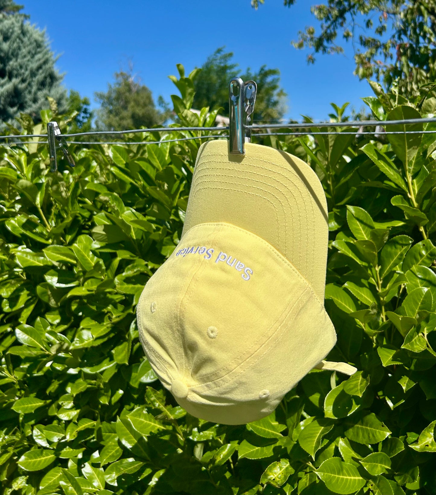 Light yellow cap with embroidered text ‘Sand Service’, hanging on a line against a bright green hedge and clear blue sky