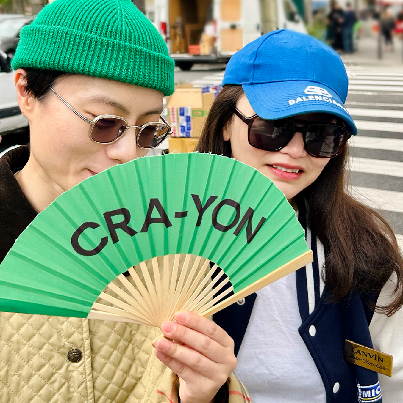 Two stylish people holding a green CRA-YON fan on a city street, wearing sunglasses and colorful caps.
