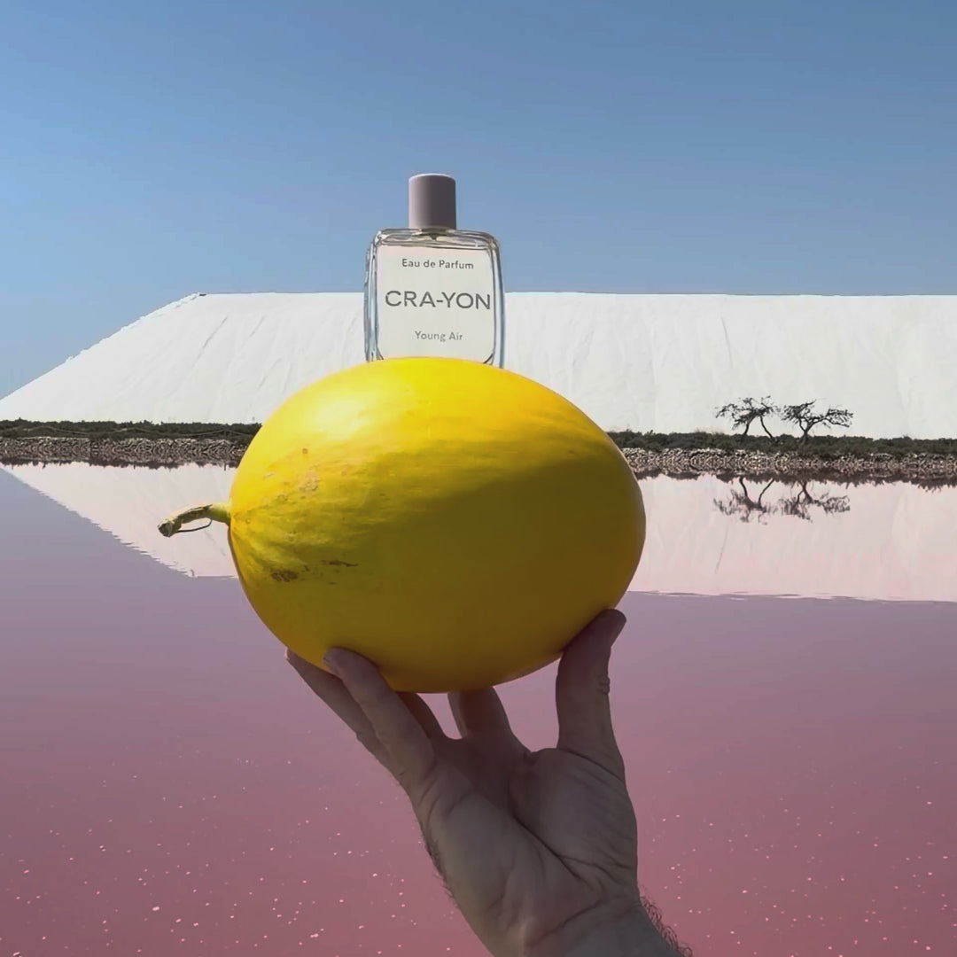 CRA-YON Young Air Eau de Parfum bottle balanced on a yellow melon against a surreal landscape of blue sky, white mountains and pink water.