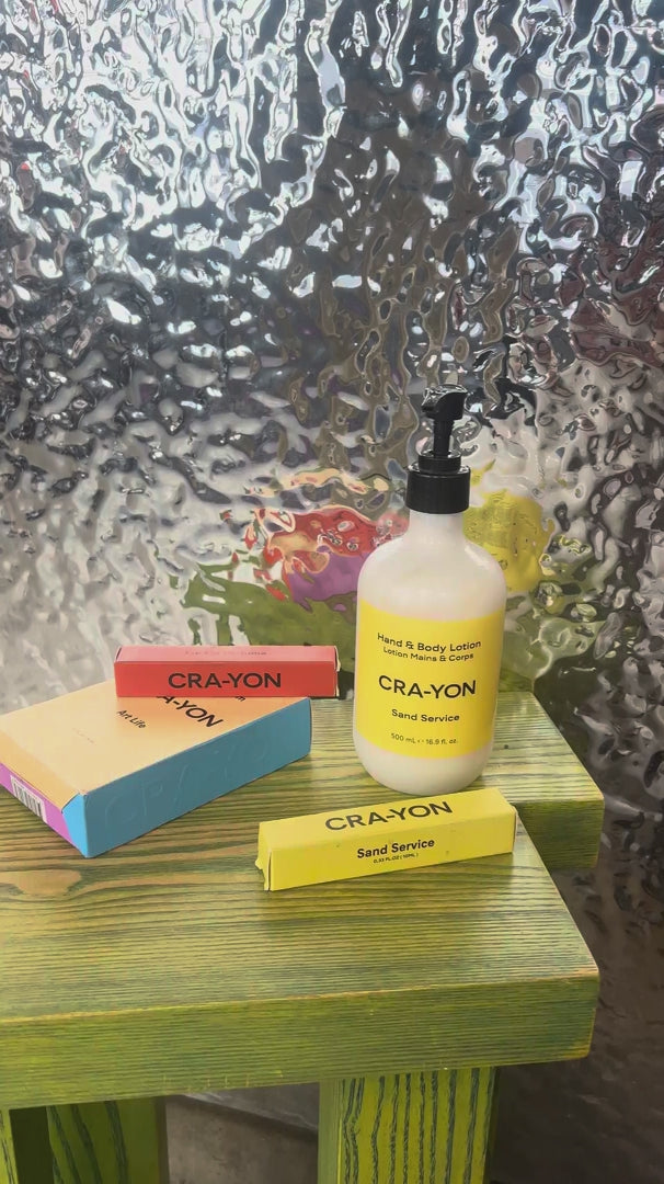 CRA-YON body lotion bottle with pump placed on a green wooden surface, alongside colorful perfume boxes, set against a textured, translucent glass background with soft light reflections.