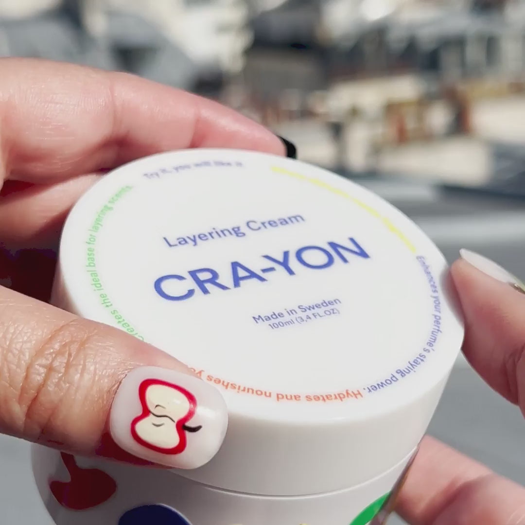 CRA-YON Layering Cream jar held in hand with close-up view of product packaging. Skincare and fragrance layering cream made in Sweden, shown with colorful branding and creative nail art design. Urban city background blurred in sunlight.